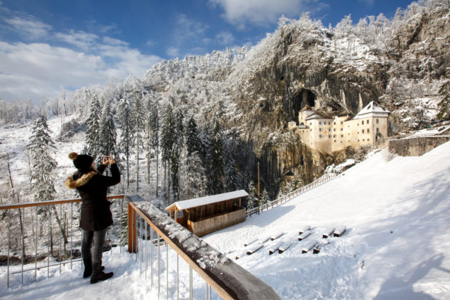 A woman photographing Predjama Castle blanketed with snow in winter